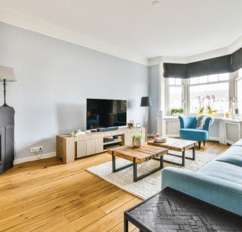 A bright, modern living room featuring light wood flooring, a light blue sofa, nested wooden coffee tables on a geometric rug, a flat-screen TV, and a blue armchair situated next to a large window.