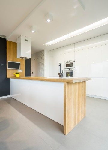 Here are a few alt text options for this image, ranging from most detailed to brief: Option 1 (Descriptive & SEO friendly): "A bright, modern minimalist kitchen featuring a long island with a light wood base and white countertop, white cabinetry with built-in stainless steel appliances, dark grey accent cabinets, and light grey flooring.